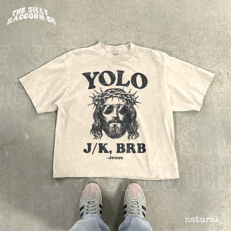 A white t-shirt with a graphic design of a man wearing sunglasses and a crown, along with the text "YOLO J.K. BRB Jesus" printed on it. The t-shirt is laid out on a concrete floor, and a pair of sneakers is visible underneath it.