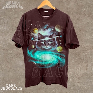 A dark t-shirt with a graphic design depicting a cat surrounded by a swirling galaxy and celestial objects, including planets and stars.