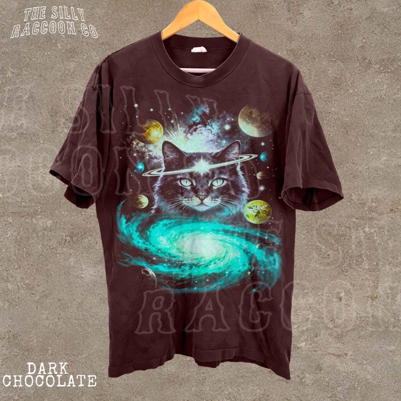 A dark t-shirt with a graphic design depicting a cat surrounded by a swirling galaxy and celestial objects, including planets and stars.