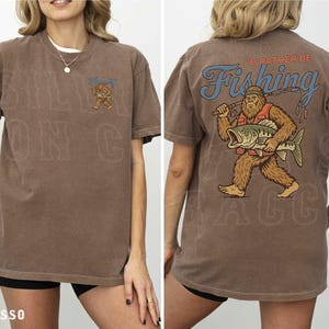 a woman wearing a brown t-shirt with a graphic design on the back. The design depicts a large fish with a bear-like appearance, and the text "I'D Rather Be Fishing" is written above the fish.