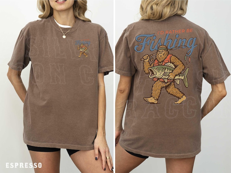 a woman wearing a brown t-shirt with a graphic design on the back. The design depicts a large fish with a bear-like appearance, and the text "I'D Rather Be Fishing" is written above the fish.