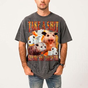 A man wearing a gray t-shirt with a graphic design featuring a dog and toilet paper rolls, along with the text "TAKE A SHIT BEFORE SHIT TAKES YOU".