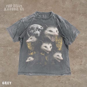 A grey t-shirt with a graphic design featuring multiple opossum faces with open mouths, arranged in a stacked formation.