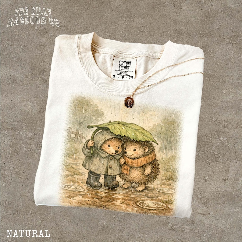 A white t-shirt with a printed illustration of two hedgehog characters standing together under an umbrella, with one hedgehog wearing a raincoat.