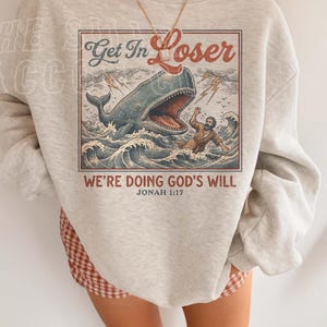 A person wearing a gray sweatshirt with a graphic design depicting a whale and a person swimming in the ocean. The text on the sweatshirt reads "Get In Loser" and "We're Doing God's Will" along with the Bible verse " Jonah 1:17".
