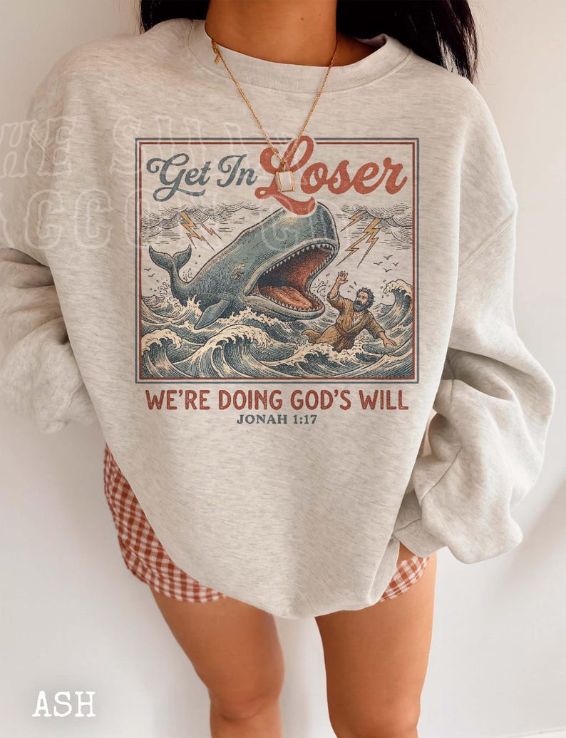 A person wearing a gray sweatshirt with a graphic design depicting a whale and a person swimming in the ocean. The text on the sweatshirt reads "Get In Loser" and "We're Doing God's Will" along with the Bible verse " Jonah 1:17".