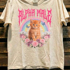 a white t-shirt with a graphic design of a cat surrounded by flowers and a rainbow.