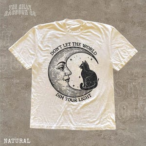 A white t-shirt with a graphic design depicting a cat sitting on a crescent moon and the text "Don't let the world dim your light" printed on it.