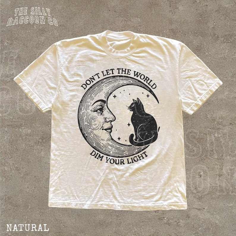 A white t-shirt with a graphic design depicting a cat sitting on a crescent moon and the text "Don't let the world dim your light" printed on it.