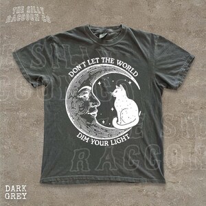 a grey t-shirt with a graphic design depicting a cat sitting on a crescent moon and the text "Don't Let the World Dim Your Light" printed on it.