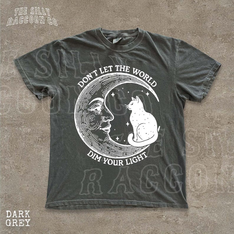 a grey t-shirt with a graphic design depicting a cat sitting on a crescent moon and the text "Don't Let the World Dim Your Light" printed on it.