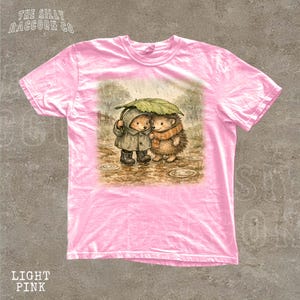 A pink t-shirt with a printed illustration of two hedgehog characters standing together under an umbrella.