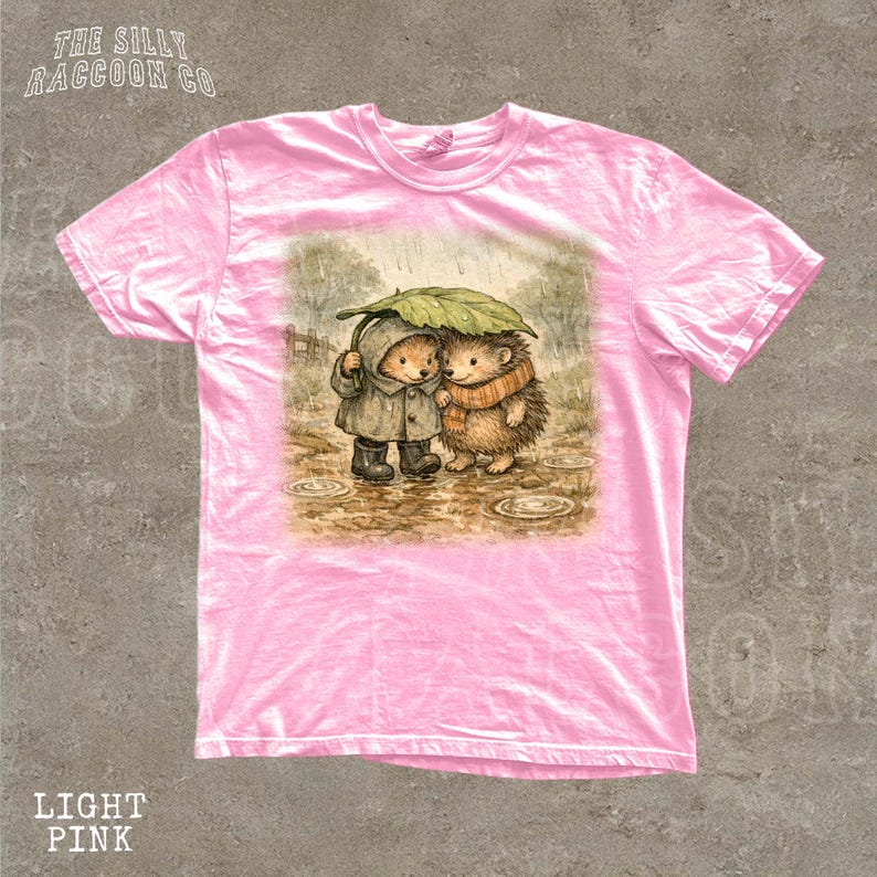 A pink t-shirt with a printed illustration of two hedgehog characters standing together under an umbrella.