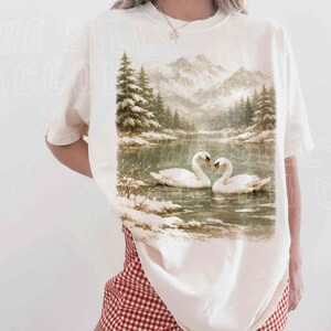 A woman wearing a white t-shirt with a printed image of two swans swimming in a lake, surrounded by snow-covered trees.