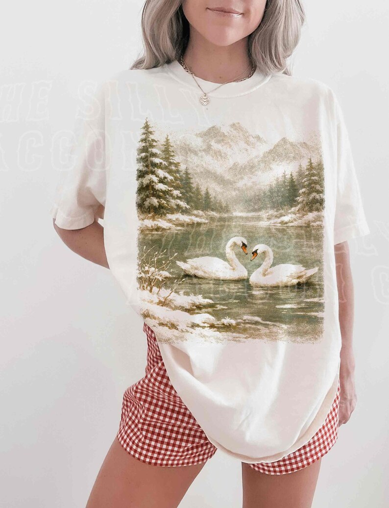 A woman wearing a white t-shirt with a printed image of two swans swimming in a lake, surrounded by snow-covered trees.