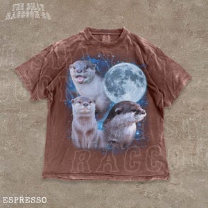 A brown t-shirt with a graphic design featuring three otters against a backdrop of a full moon and stars.