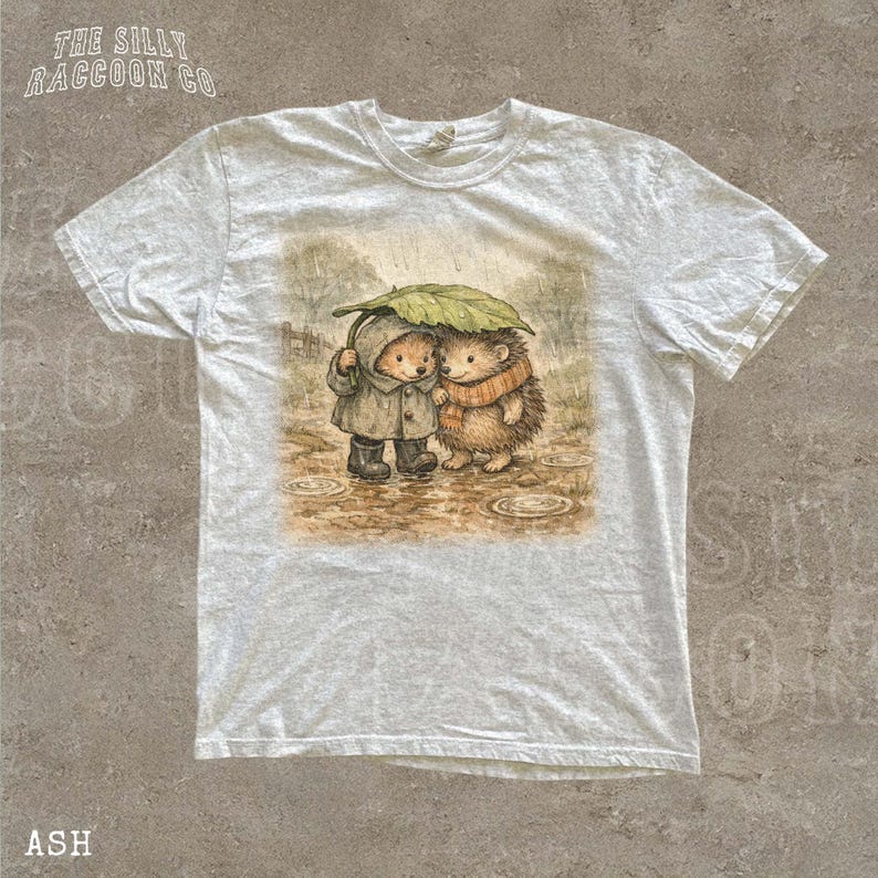 A white t-shirt with a printed illustration of two hedgehog characters standing together under an umbrella.