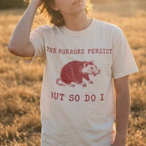 A person wearing a white t-shirt with a graphic of a red opossum and the text "The Horrors Persist BUT SO DO I" printed on it.