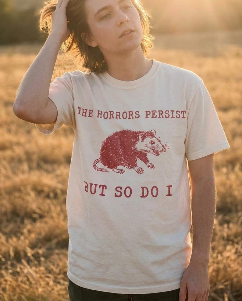 A person wearing a white t-shirt with a graphic of a red opossum and the text "The Horrors Persist BUT SO DO I" printed on it.