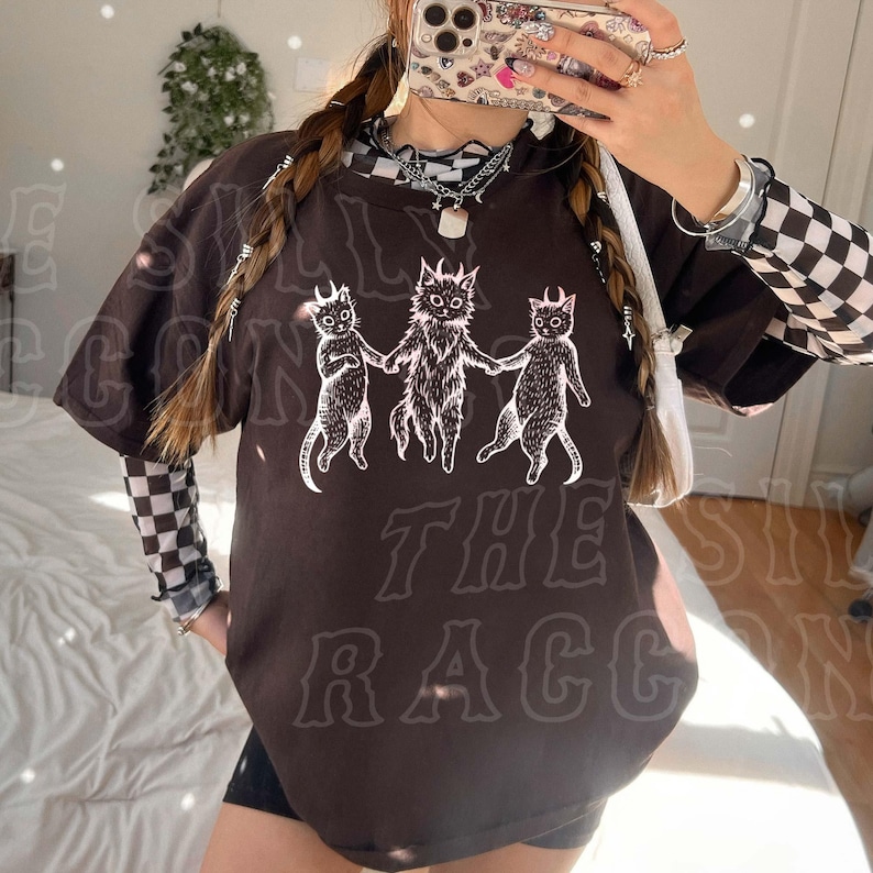 A person is taking a selfie with a black t-shirt featuring a graphic design of three cats.