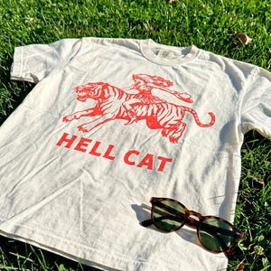 A white t-shirt with a red graphic of a tiger and the text "HELL CAT" is lying on the grass, accompanied by a pair of sunglasses.