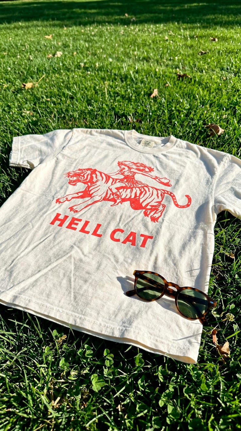 A white t-shirt with a red graphic of a tiger and the text "HELL CAT" is lying on the grass, accompanied by a pair of sunglasses.