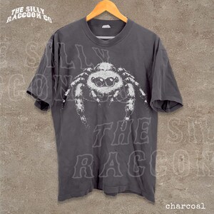 A gray t-shirt with a large white spider graphic printed on it, hanging on a wooden hanger.