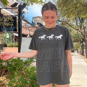 A woman wearing a black t-shirt with a graphic of three horses is standing on a sidewalk, smiling and looking down at her hand.