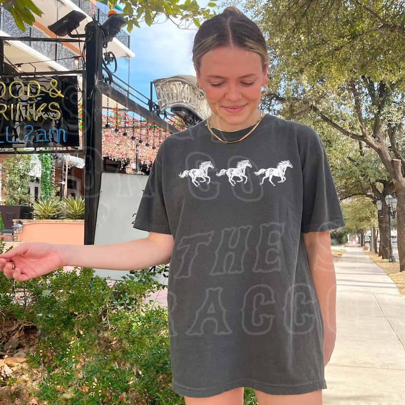 A woman wearing a black t-shirt with a graphic of three horses is standing on a sidewalk, smiling and looking down at her hand.