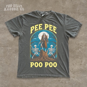 May include: Dark grey t-shirt with a graphic of a skeleton in a long coat, two skeletons with guns, and the words "PEE PEE POO POO" in yellow. The shirt has the text "THE SILLY RACCOON CO" at the top.