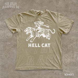 A beige t-shirt with a graphic design depicting a person riding a tiger, accompanied by the text "HELL CAT" printed on it.