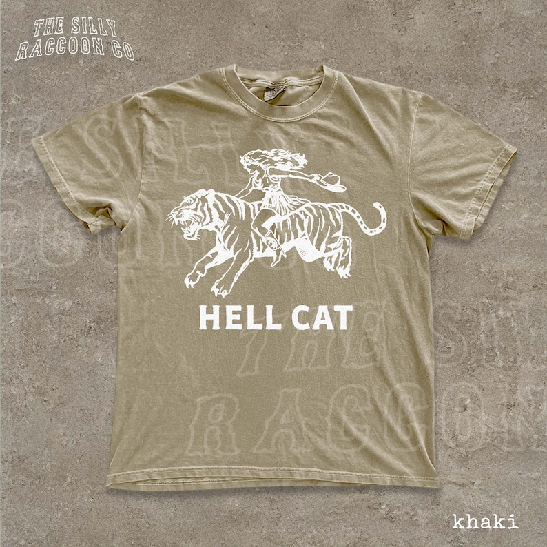 A beige t-shirt with a graphic design depicting a person riding a tiger, accompanied by the text "HELL CAT" printed on it.