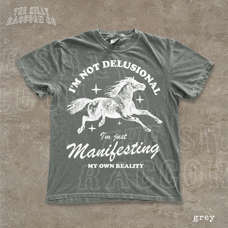 A grey t-shirt with a graphic design depicting a horse in mid-gallop and the text "I'm not delusional, I'm just Manifesting, MY OWN REALITY" printed on it.