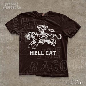 A dark brown t-shirt with a graphic design depicting a woman riding a tiger, accompanied by the text "HELL CAT" and "dark chocolate".