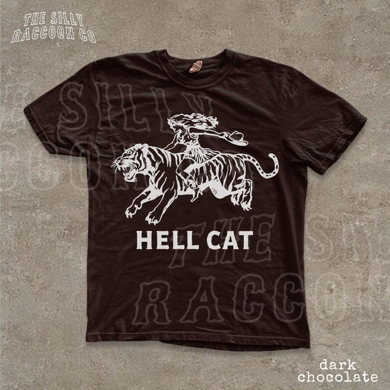 A dark brown t-shirt with a graphic design depicting a woman riding a tiger, accompanied by the text "HELL CAT" and "dark chocolate".