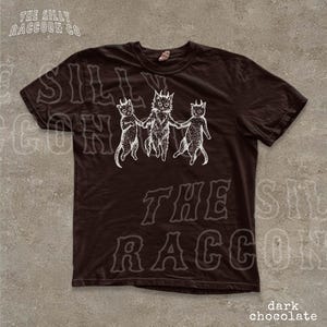 A dark brown t-shirt with a graphic design featuring three cats with horns and a raccoon-like creature. The text on the t-shirt reads "SILLY ON THE RACCOON CO. dark chocolate".