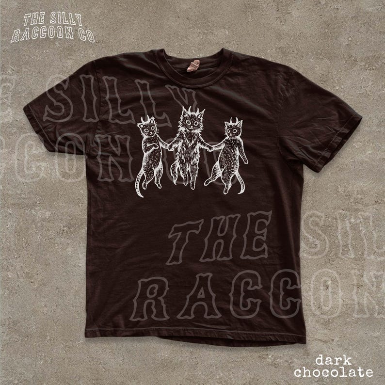 A dark brown t-shirt with a graphic design featuring three cats with horns and a raccoon-like creature. The text on the t-shirt reads "SILLY ON THE RACCOON CO. dark chocolate".