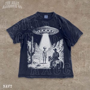 The image is a t-shirt design featuring a scene with an alien spacecraft hovering above a desert landscape, where a man on a horse is present.