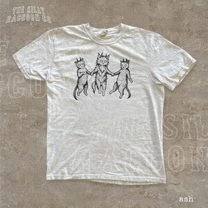 A white t-shirt with a graphic design featuring three cats with horns, depicted in a whimsical and playful manner.