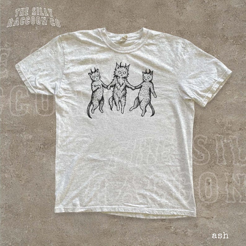 A white t-shirt with a graphic design featuring three cats with horns, depicted in a whimsical and playful manner.