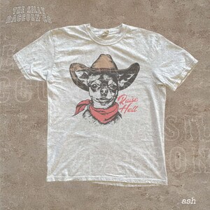 A white t-shirt with a graphic design of a dog wearing a cowboy hat and a red scarf, along with the text "Raise Hell" printed on it.