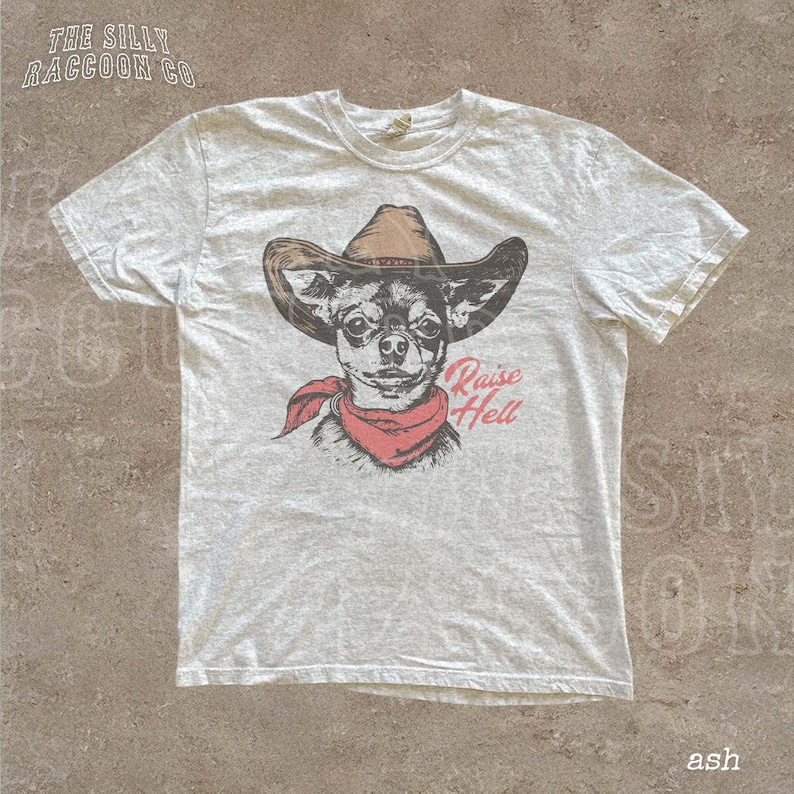 A white t-shirt with a graphic design of a dog wearing a cowboy hat and a red scarf, along with the text "Raise Hell" printed on it.