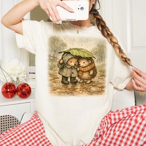 A person is taking a selfie with a white camera, wearing a white t-shirt with a printed illustration of two hedgehog characters sharing an umbrella.