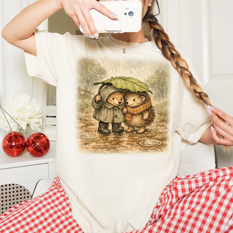 A person is taking a selfie with a white camera, wearing a white t-shirt with a printed illustration of two hedgehog characters sharing an umbrella.