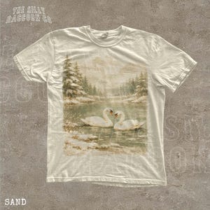 A white t-shirt with a printed image of two swans swimming in a lake, surrounded by snow-covered trees.