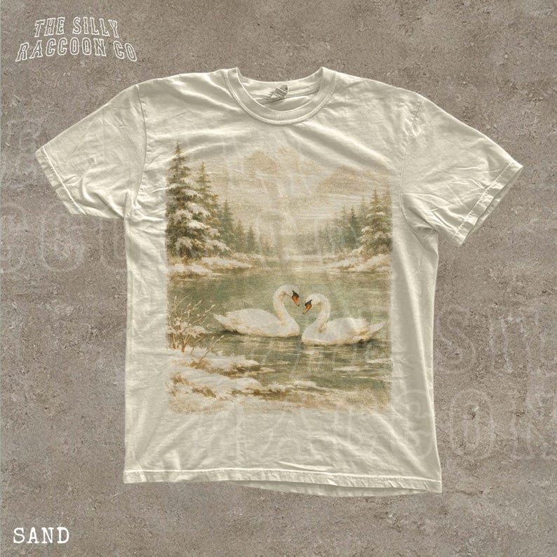 A white t-shirt with a printed image of two swans swimming in a lake, surrounded by snow-covered trees.