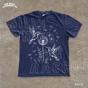 A navy blue t-shirt with a graphic design featuring two hummingbirds and dandelions against a dark background.