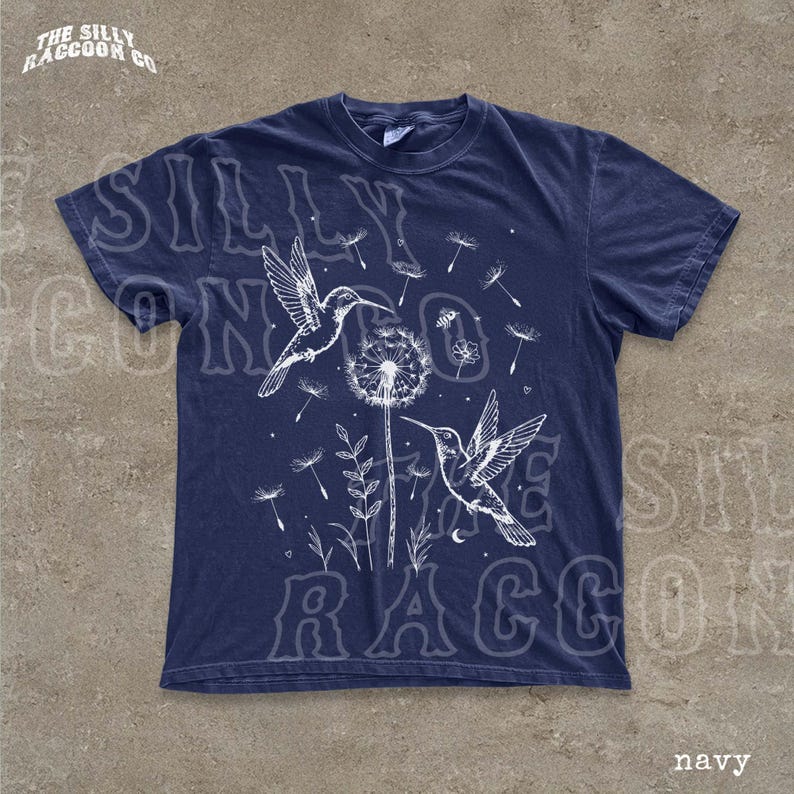 A navy blue t-shirt with a graphic design featuring two hummingbirds and dandelions against a dark background.