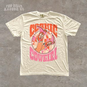 A white t-shirt with a graphic design featuring a pair of boots and the text "COSMIC COWEGIRL" in a retro style.