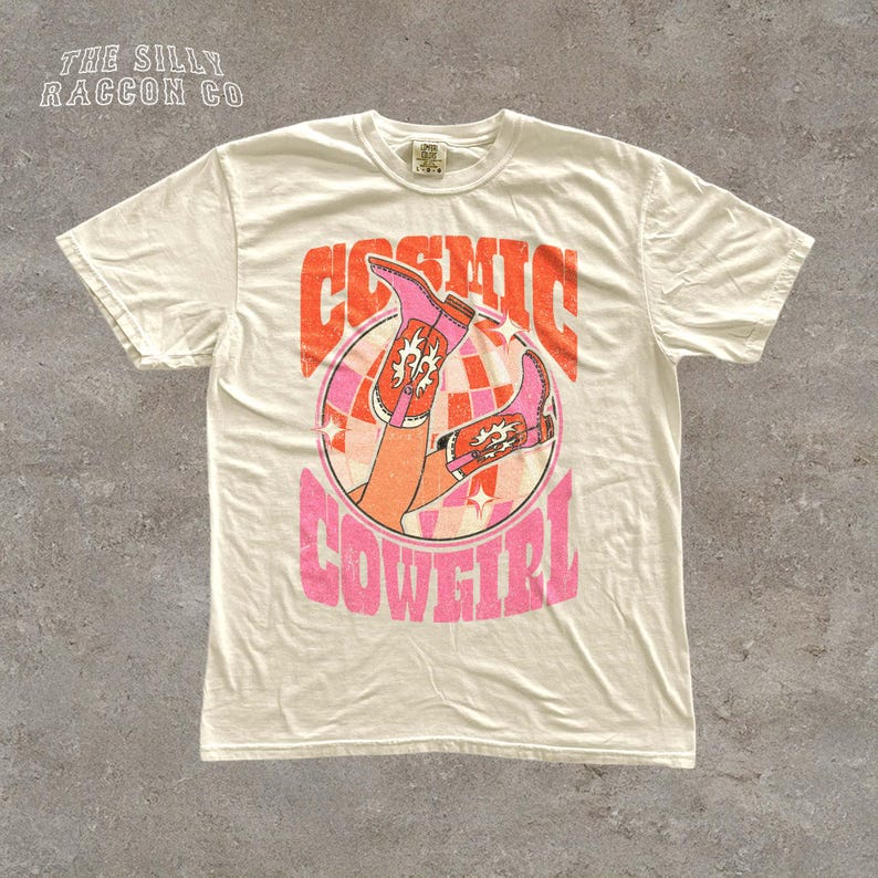 A white t-shirt with a graphic design featuring a pair of boots and the text "COSMIC COWEGIRL" in a retro style.